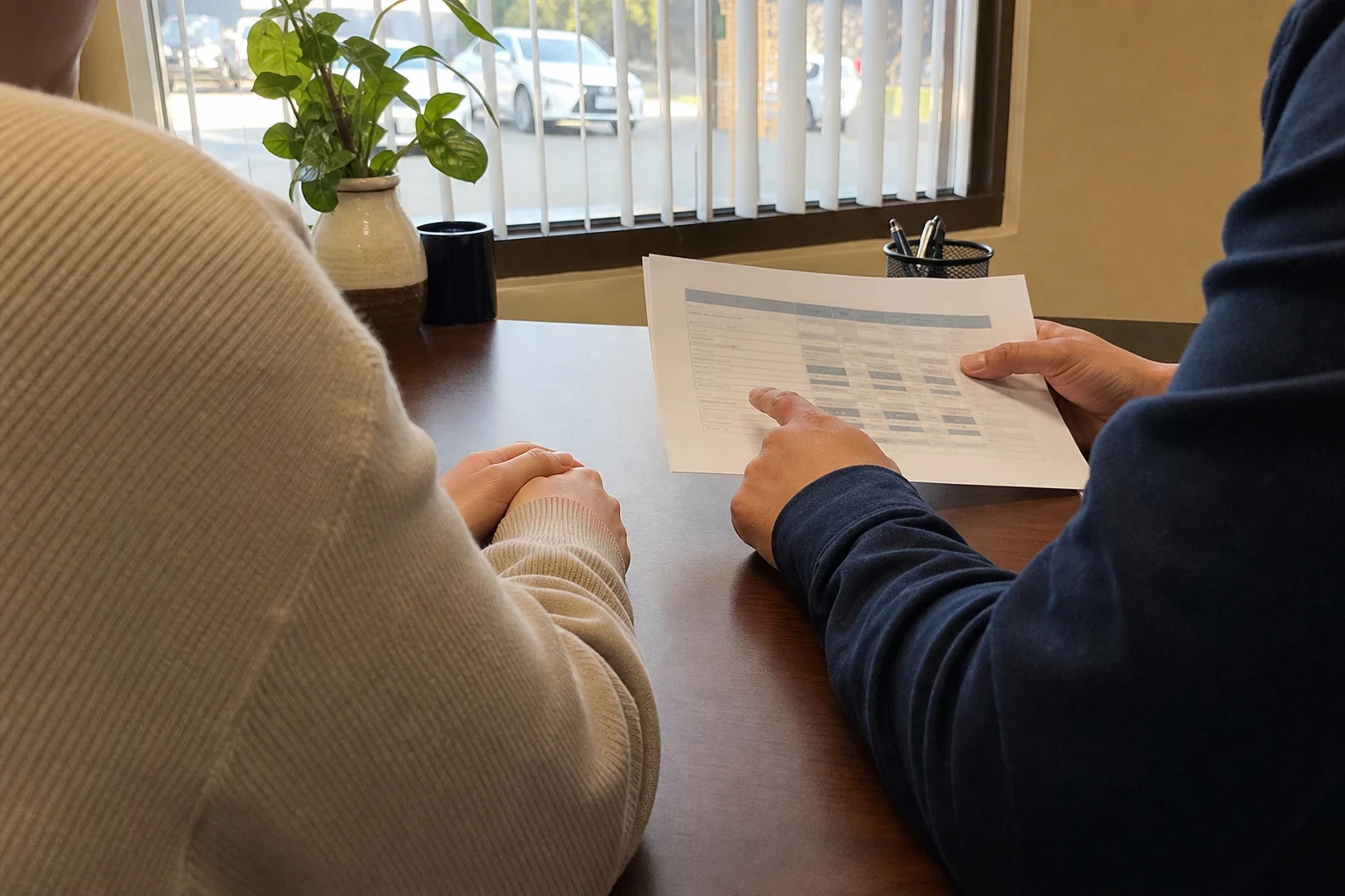 Over-the-shoulder view of an agent pointing to a printed comparison sheet for a client at an office desk by a window with blinds and a potted plant
