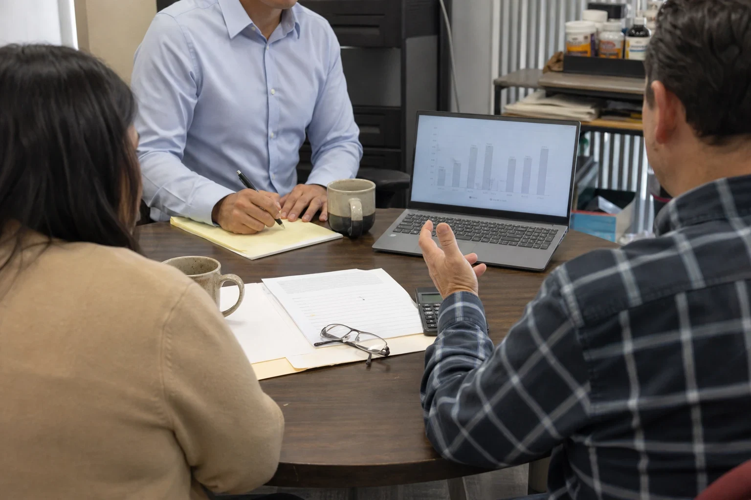 Agent in a light blue button-down taking notes on a legal pad at a round office table while a couple talks, with a laptop showing a bar chart and papers nearby